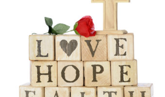 Rustic wood alphabet blocks arranged to say LOVE, HOPE and FAITH. They're topped with a red rose and a wood cross.