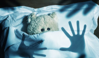 An adorable teddy bear laying in bed, scared by the shadow of human hands, under the sheets.